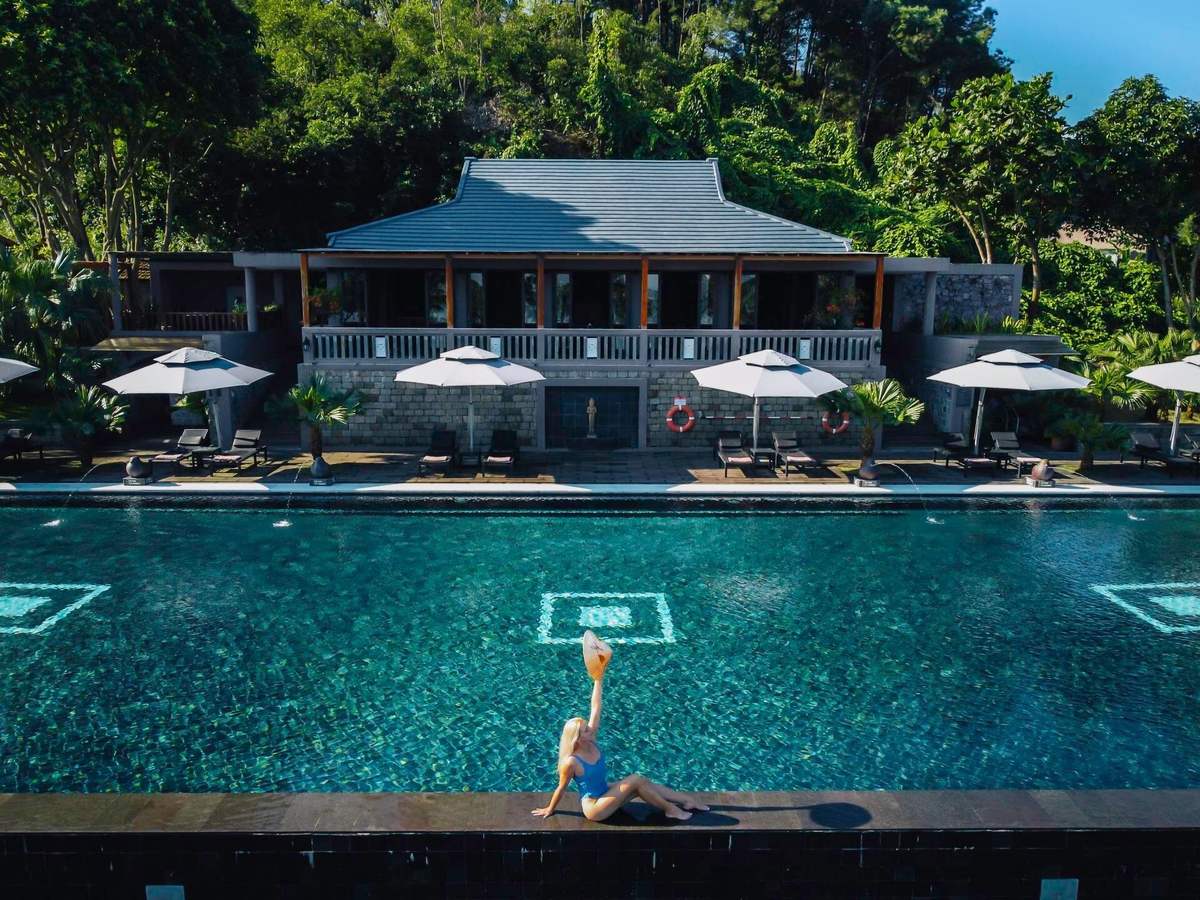 Long teal pool with white diamond tile motifs, a woman in a swimsuit sitting on its edge, white umbrellas and a gray-roofed pavilion against green forest