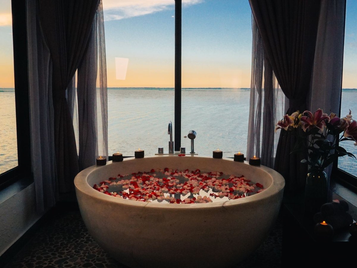 Round stone bathtub filled with red and white petals and ringed by candles, in front of tall windows over calm water at dusk