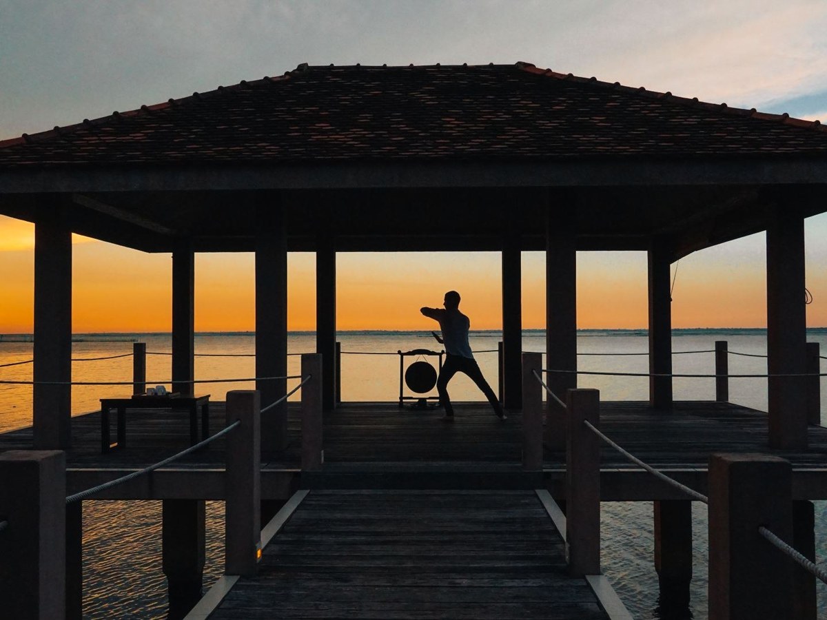 Silhouette of a person practicing tai chi under a tiled pavilion on a jetty, with an orange sky over the water behind