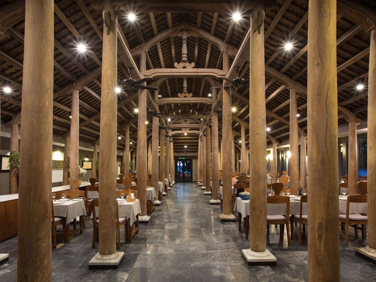 Long dining hall lined with tall round wooden columns under a carved timber roof, white-clothed tables on either side