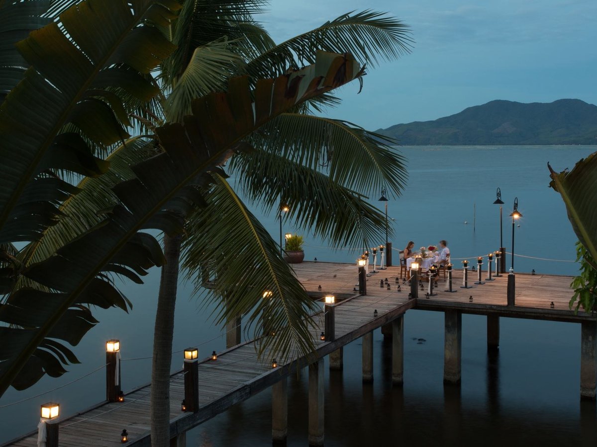Wooden jetty lined with small lamps at dusk, with a few people seated at a table at its end and dark hills across the water