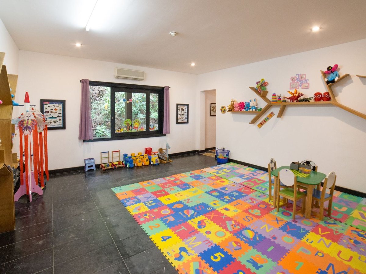 Bright playroom with a colorful foam alphabet floor, a small table and chairs, toy shelves shaped like branches and a window onto trees