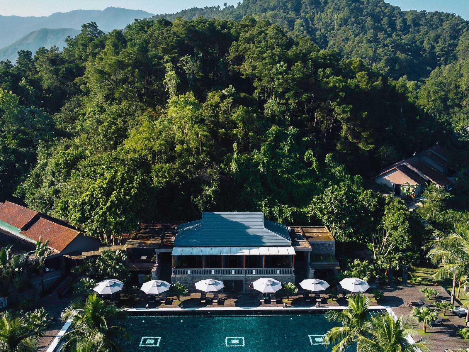 Aerial view of a long dark-tiled pool lined with white umbrellas in front of a gray-roofed pavilion, with a forested hill rising behind