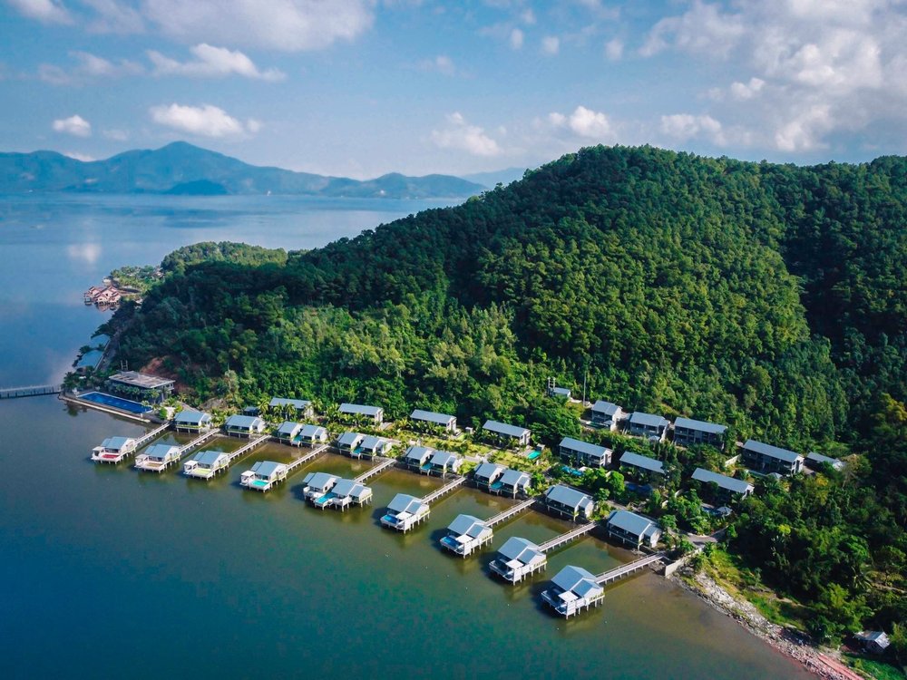 Aerial view of a forested headland with a long row of villas on stilts linked to the shore by wooden jetties, mountains across the lagoon