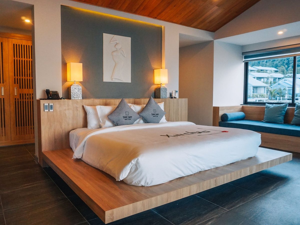 Bedroom with a low wooden platform bed, white linen, gray cushions, a timber ceiling and a window seat looking out on villas