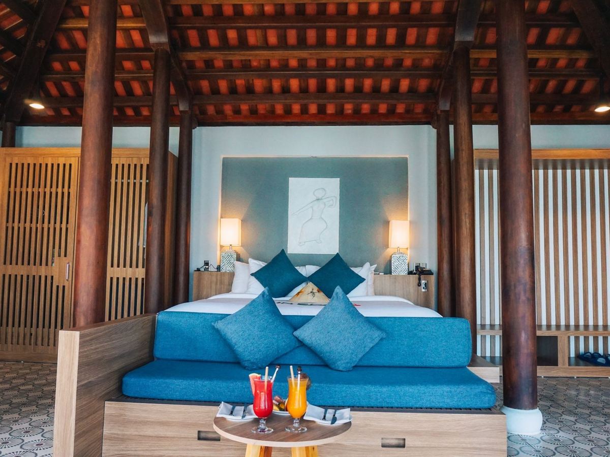 Bed with blue cushions framed by dark wooden columns under an exposed timber roof, two glasses of juice on a small round table