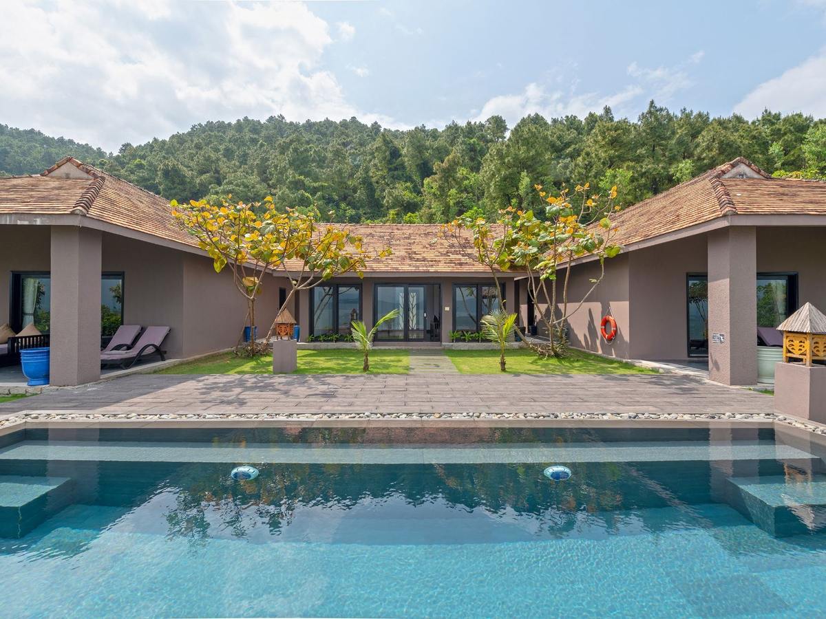 Long pool in front of a single-story villa with a tiled roof, a lawn courtyard, sun loungers and a forested hill behind