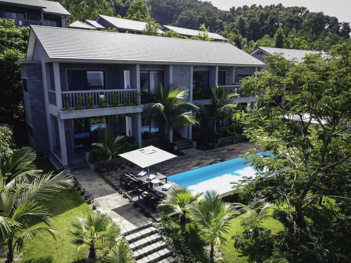 Two-story gray villa with balconies above a pool and a white parasol, stone steps climbing through a palm garden