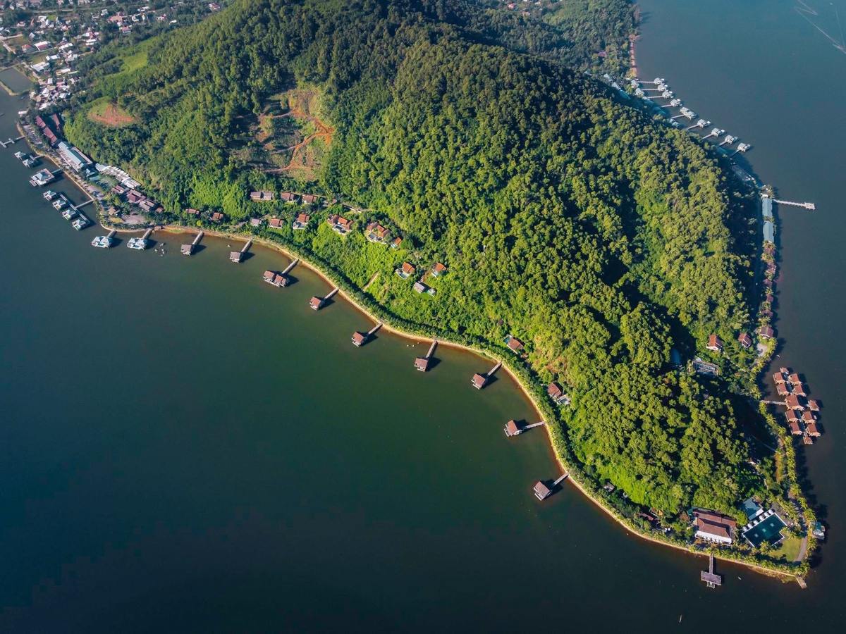 Aerial view of a forested headland curving into a wide lagoon, villas on stilts along its shore and a village at the far end