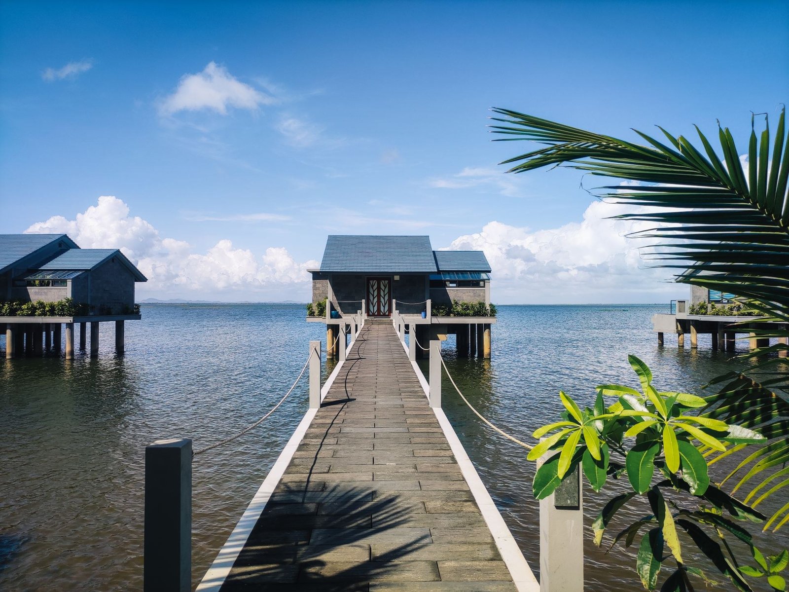 Long walkway with rope railings leading across calm water to an over-water villa with a slate-gray roof under a blue sky