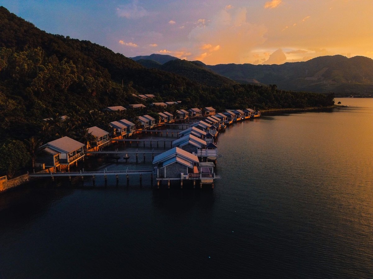Row of over-water villas on stilts lit by an orange sunset, a forested slope behind and still water in front