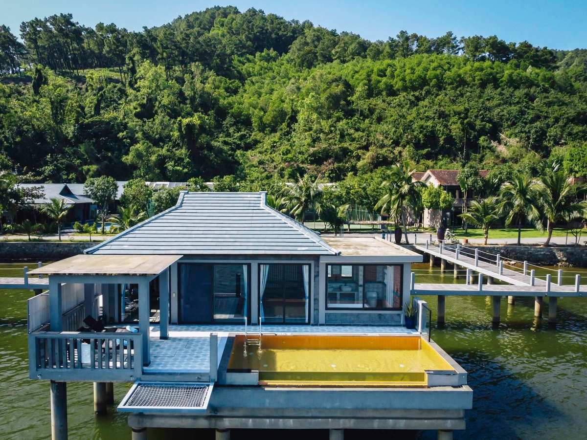 Over-water villa with a gray tiled roof and a rectangular pool on its deck, palm trees and a green hill behind