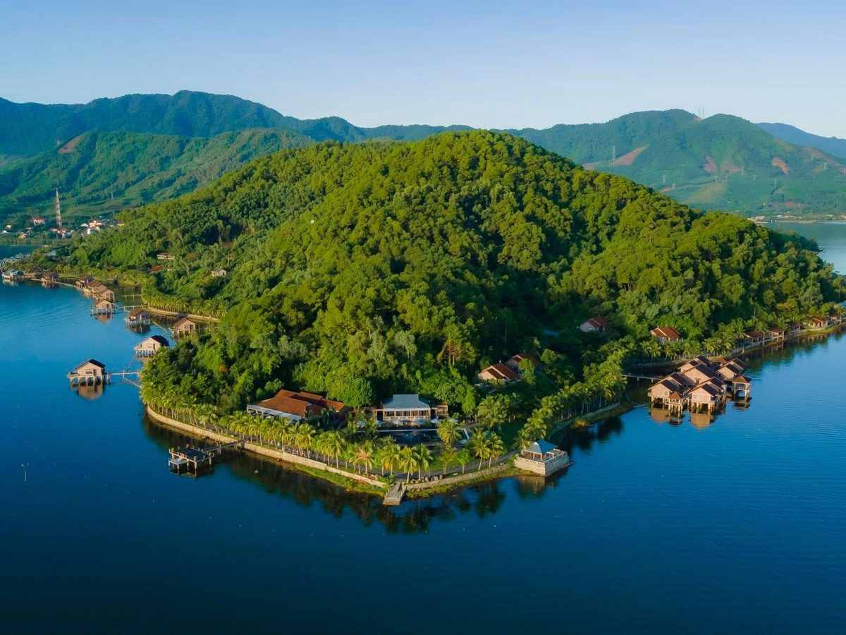 Aerial view of a wooded headland ringed by villas on stilts, with blue water around it and green mountains in the background