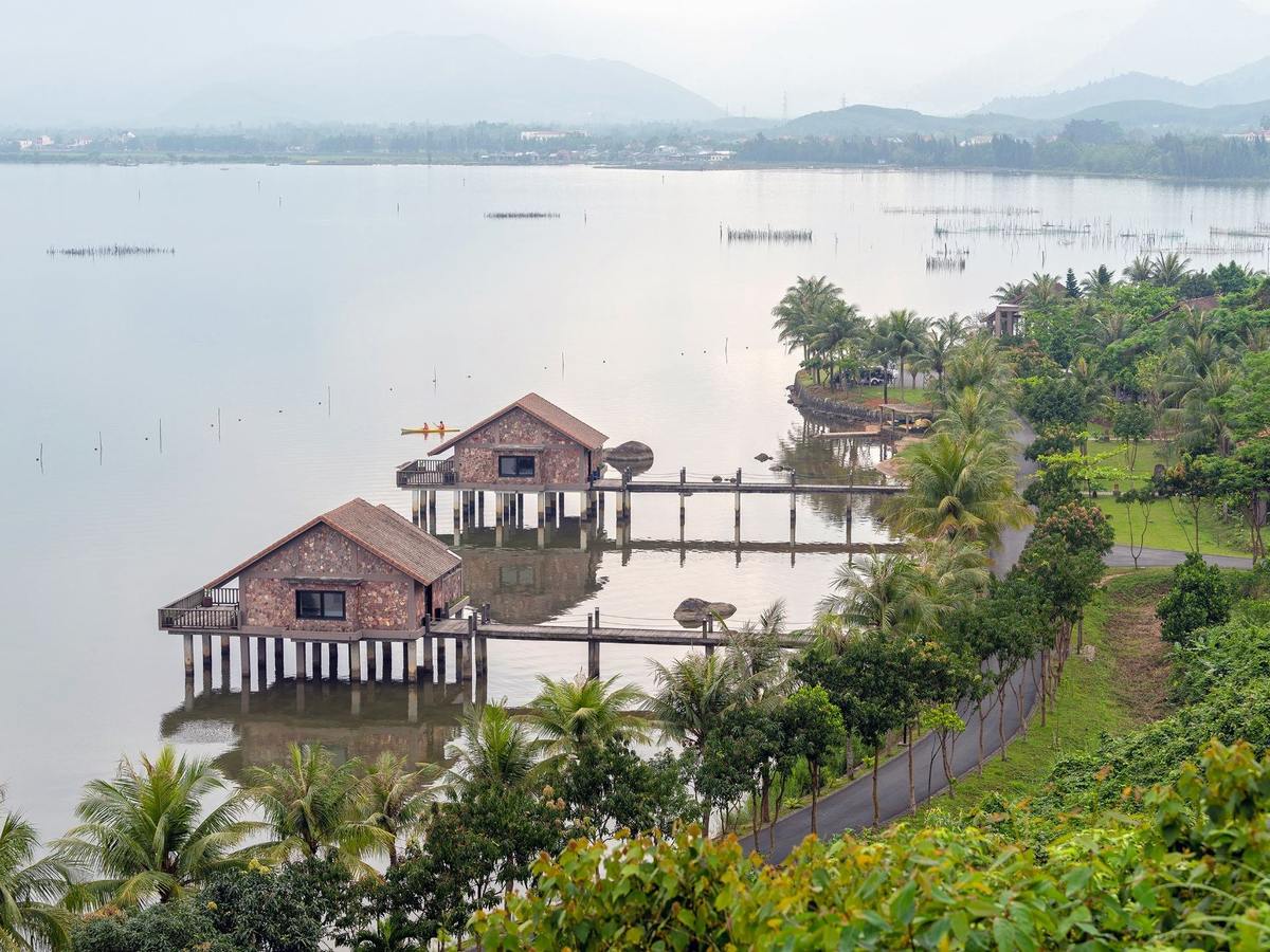 Two brick villas with red-brown roofs standing on stilts in a pale misty lagoon, fishing stakes in the water and palms along a road