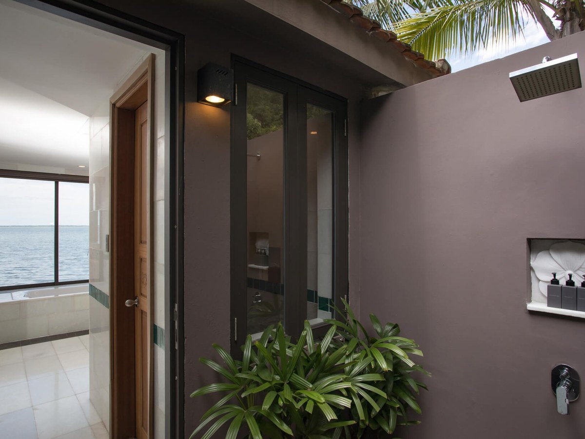 Walled outdoor shower with a rain head and a carved niche of toiletries beside a bathroom whose window looks onto the water