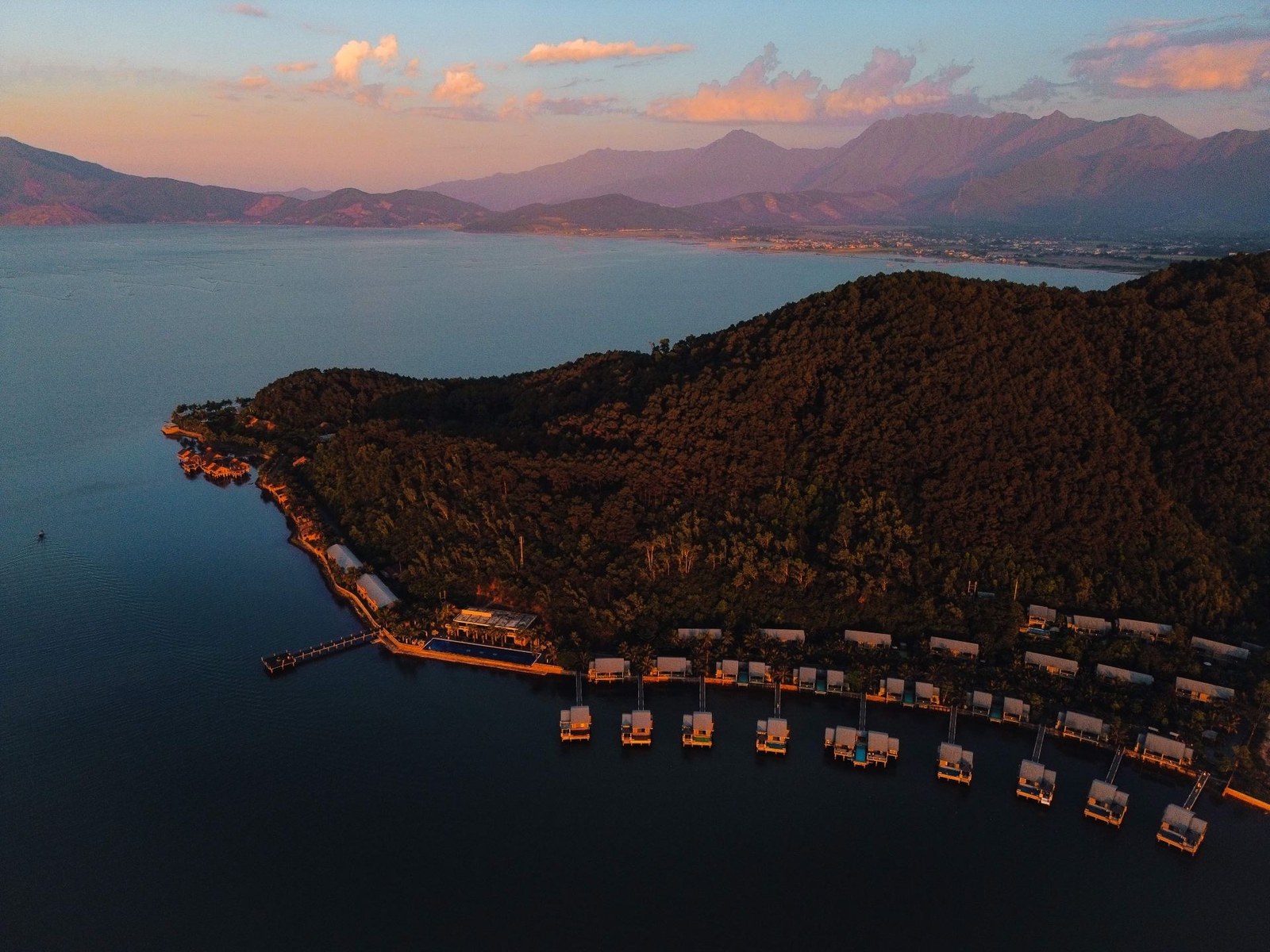 Aerial view at dusk of a forested headland with a curved line of over-water villas along its shore and pink-lit mountains across the water
