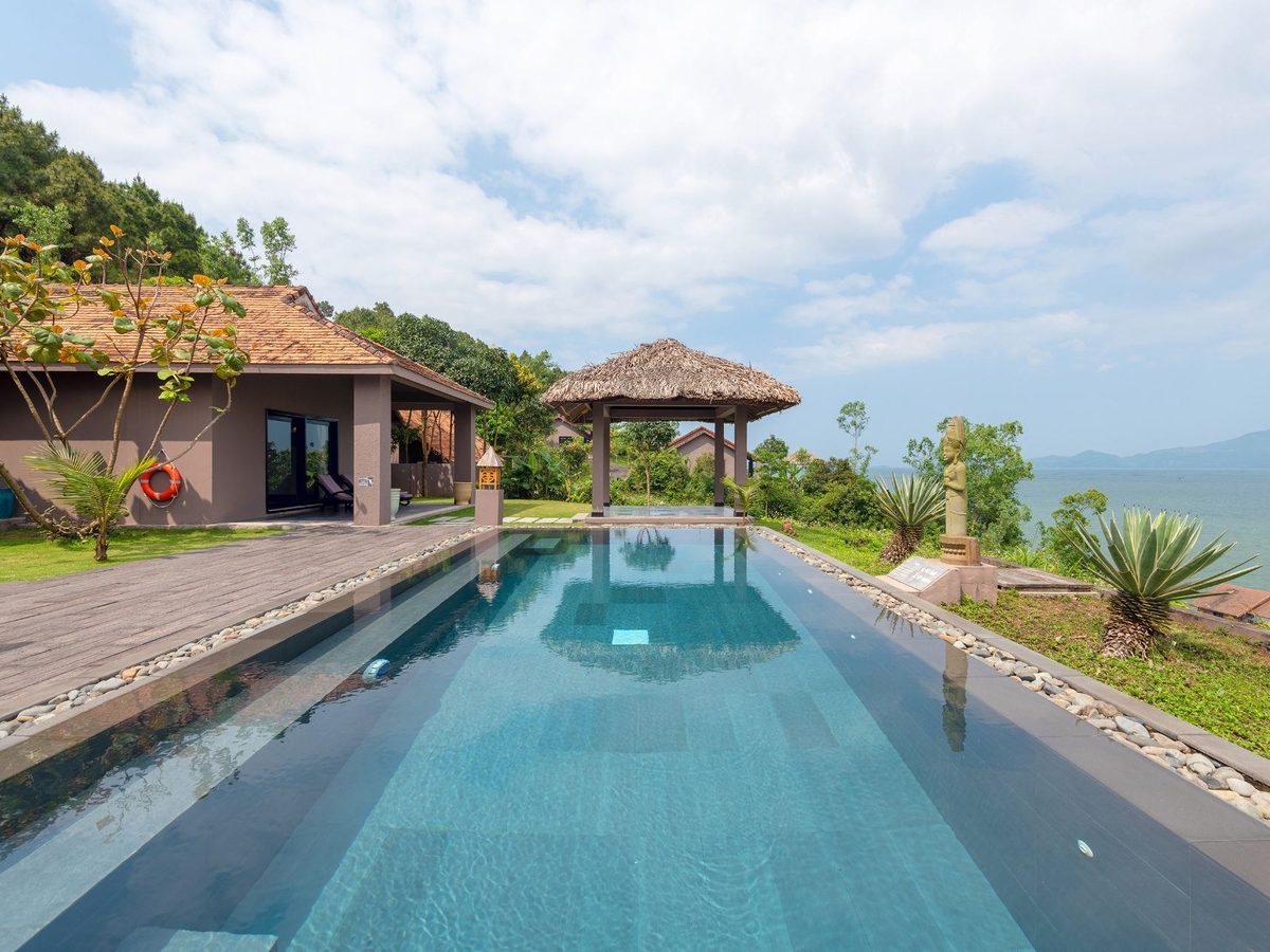 Long private pool in a lawn garden with a thatched pavilion at one end and a tiled villa behind