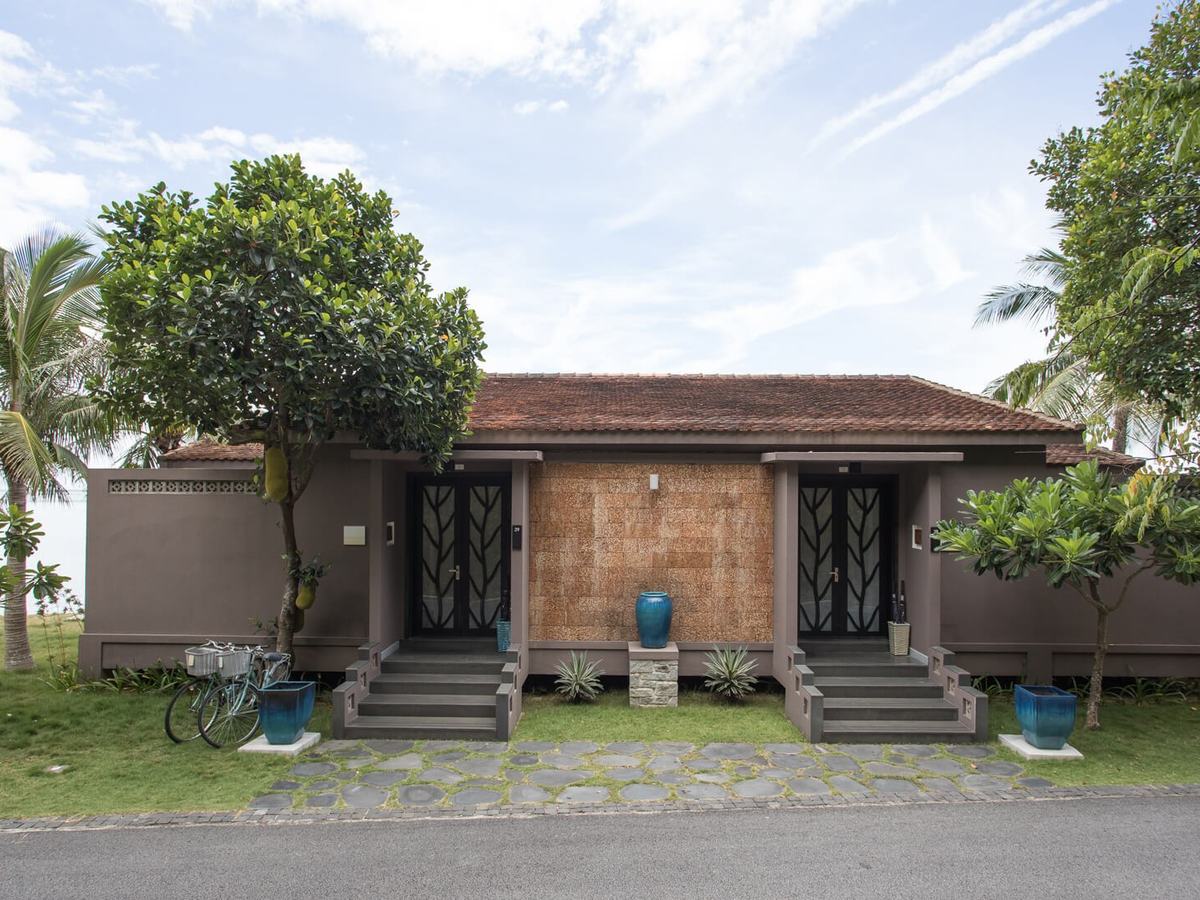 Single-story tiled villa with wooden doors and steps on a lawn under coconut palms