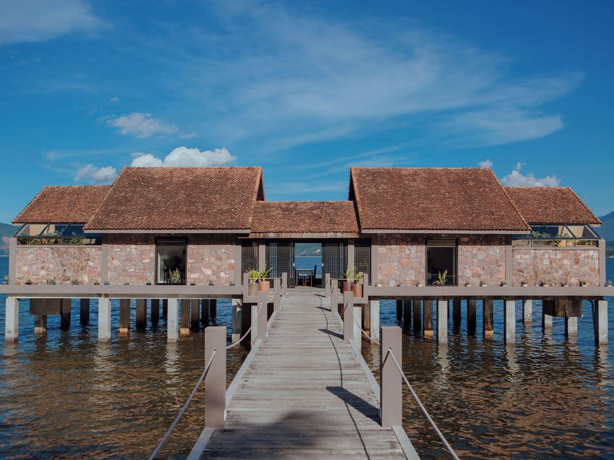 Pair of tile-roofed over-water villas linked by a walkway on stilts, blue sky and lagoon around them