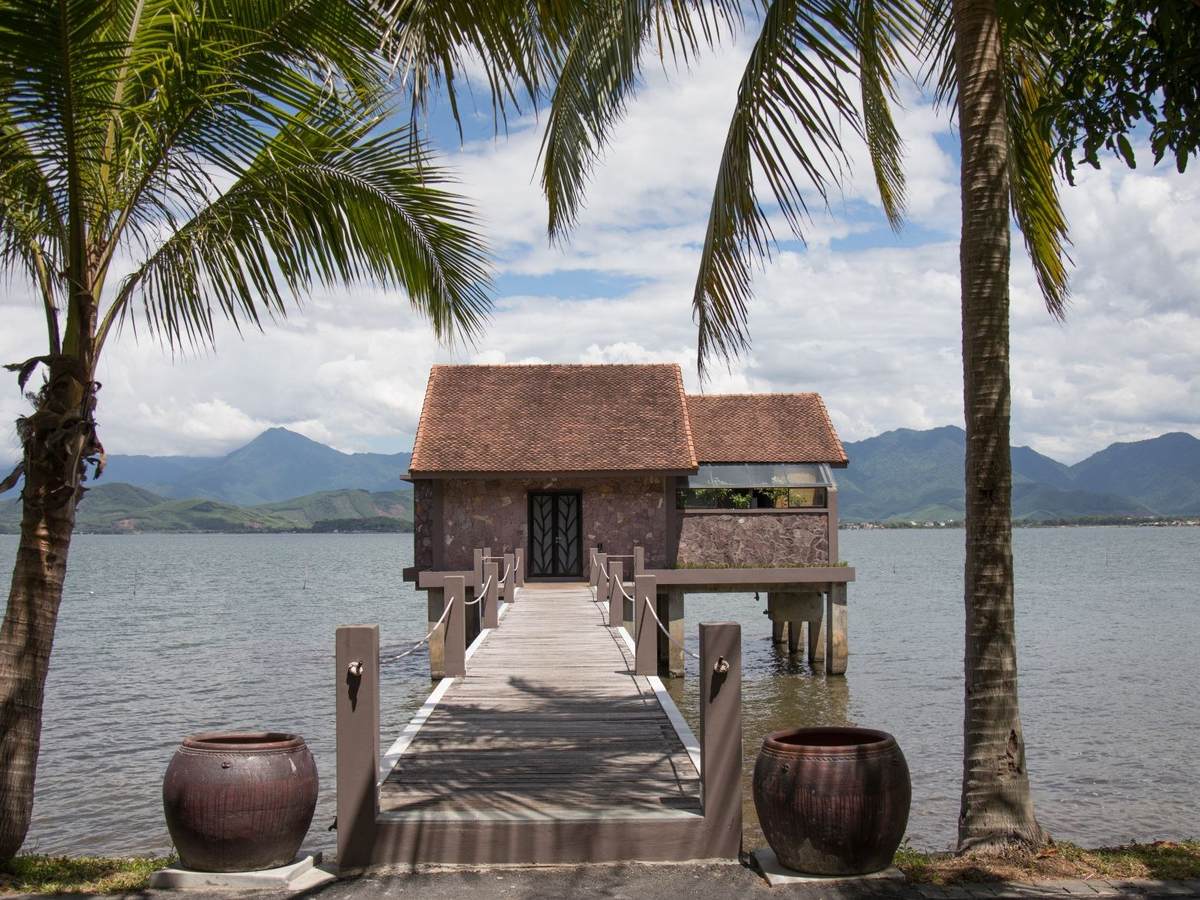Brick villa with a red-brown tiled roof standing over the lagoon at the end of a long wooden jetty between palms