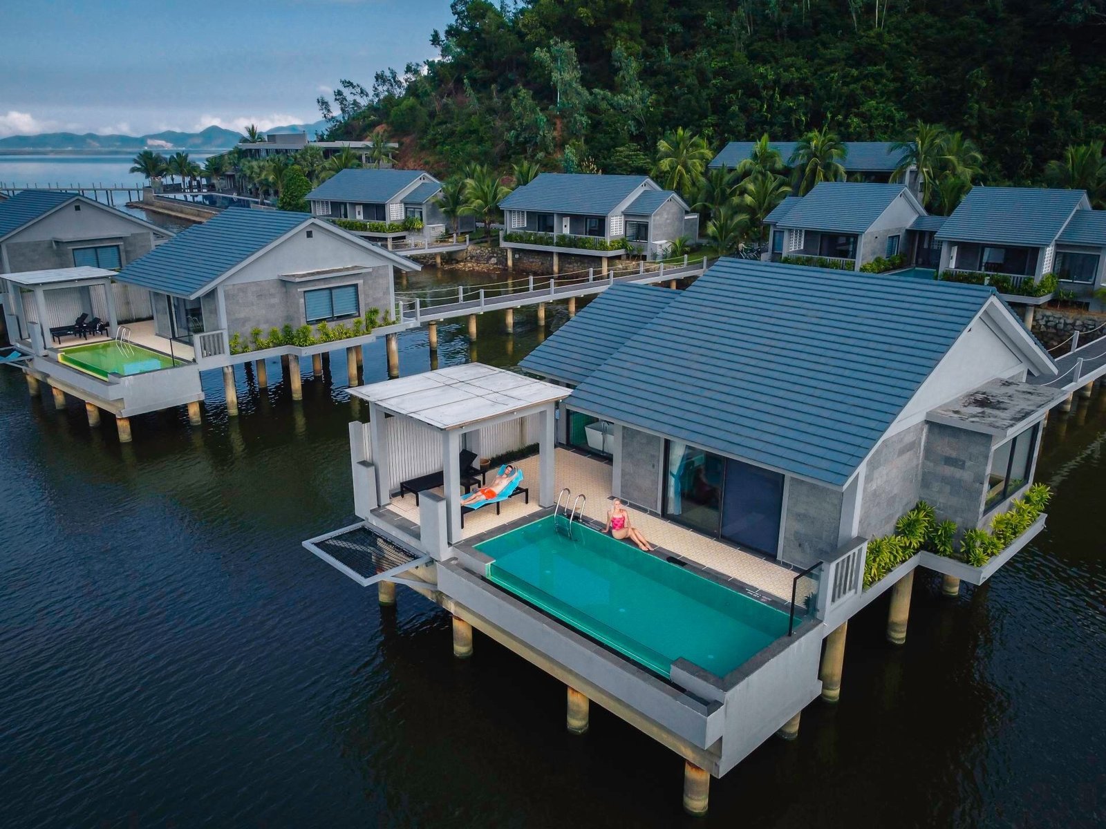 Row of over-water villas with slate-grey roofs and private pools on decks, joined to a palm-lined shore by timber walkways