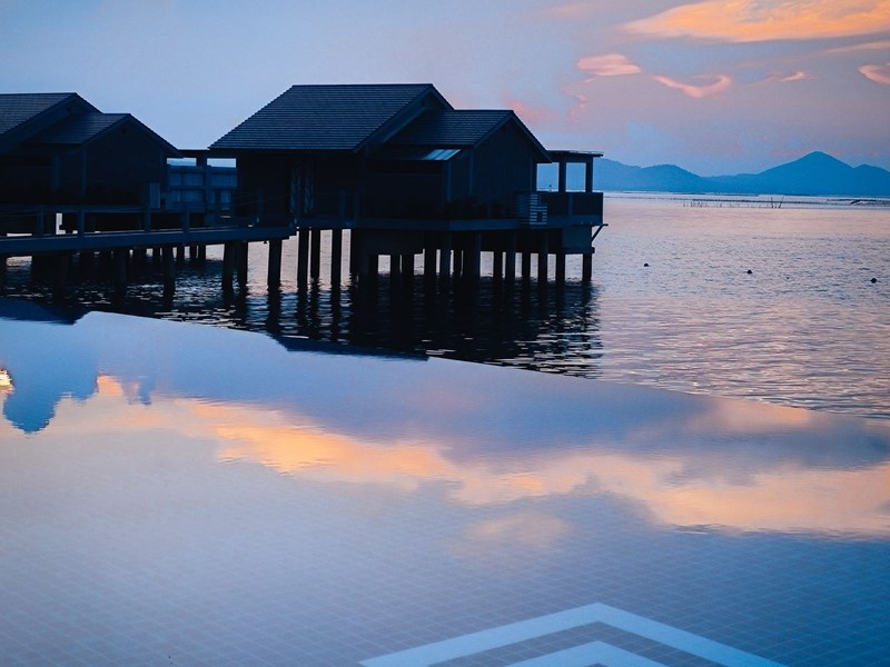 Long infinity pool at dusk reflecting villas on stilts and a pink sky over the lagoon
